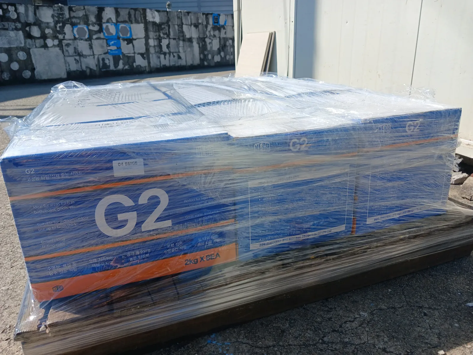 MEGA G2 줄눈시멘트 04 베이지 컬러로 표기된 박스 팔레트가 시멘트 벽돌 벽 앞에 놓인 모습